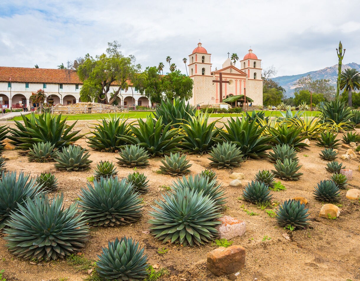 Historic Old Mission Santa Barbara with twin bell towers, red-tiled roof, and surrounding desert garden filled with agave and succulents.