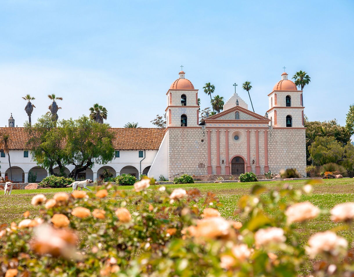 Old Mission Santa Barbara with twin bell towers, red-tiled roof, and rose garden in the foreground under a clear blue sky.