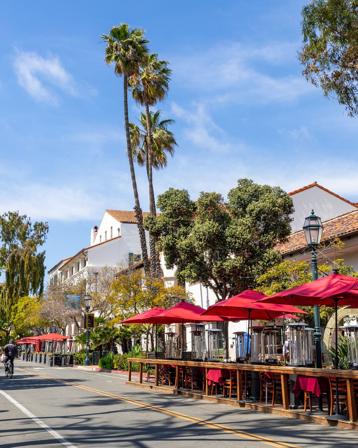 State Street in Santa Barbara with red café umbrellas, palm trees, and pedestrians walking and biking under a clear blue sky.