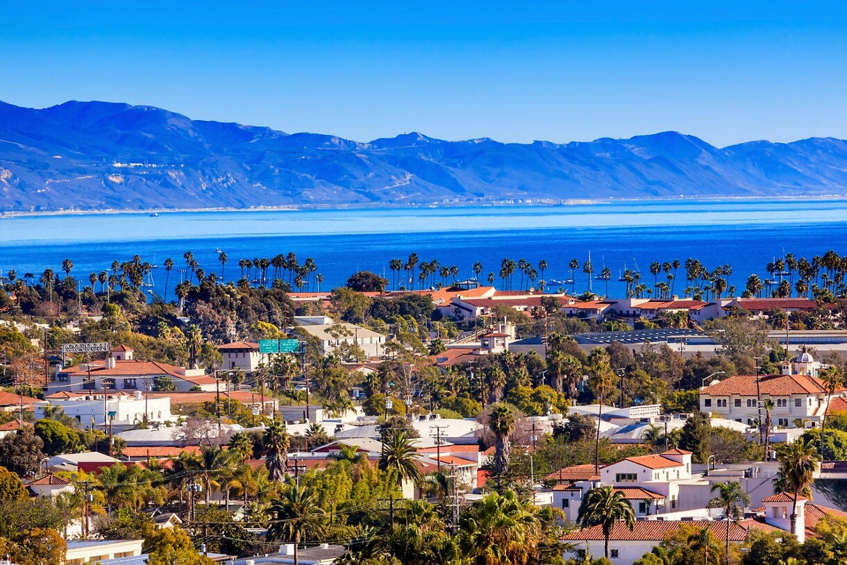 Aerial view of Santa Barbara with red-roofed buildings, palm trees, and the Pacific Ocean backed by distant mountains.
