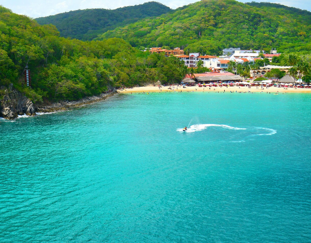 Santa Cruz Bay in Huatulco with turquoise water, jet ski trails and a sandy beach lined with palm trees, restaurants and colorful buildings against green hills.
