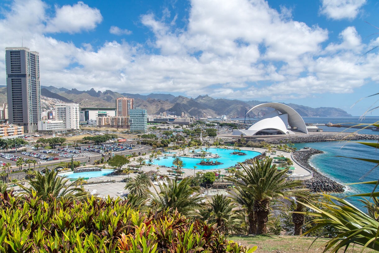 Aerial view of Santa Cruz de Tenerife with the Auditorio de Tenerife, turquoise lagoons, palm trees and mountains in the background along the Canary Islands shoreline.