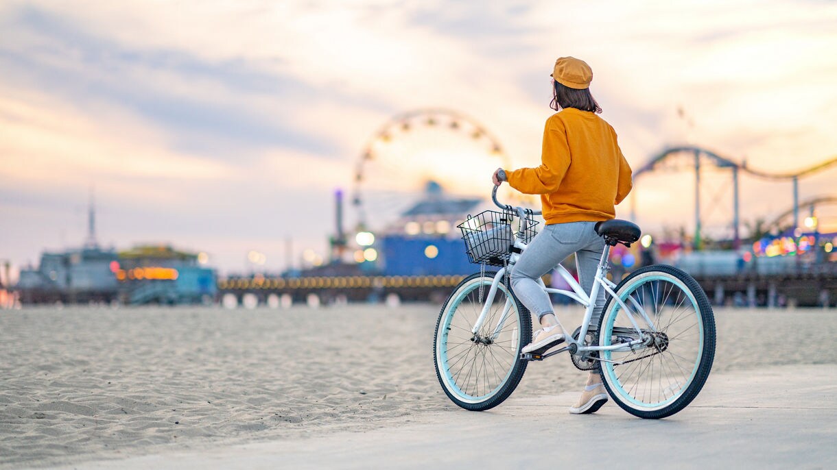 A person in an orange jacket sits on a bicycle on a beach with a Ferris wheel and amusement park pier in the background at sunset.