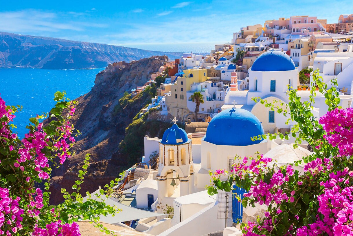 Whitewashed homes and blue-domed churches perched on a cliff overlooking deep blue sea, framed by bright pink bougainvillea.
