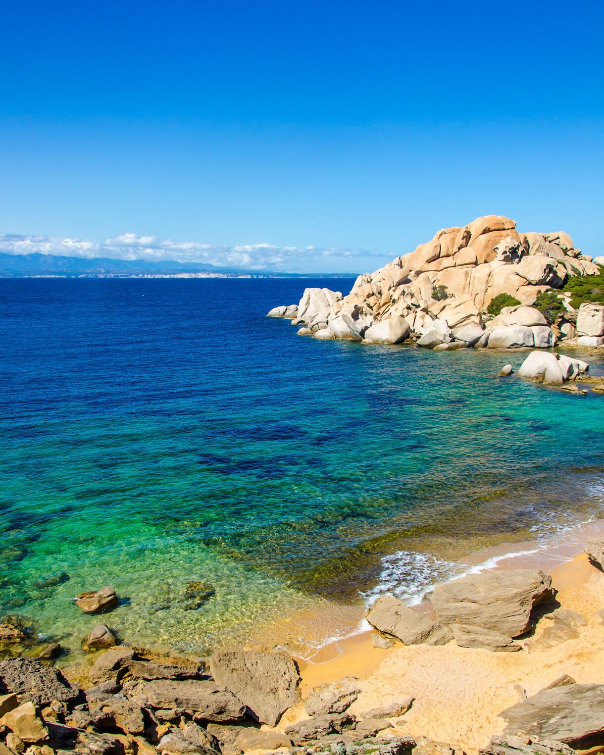 Small sandy cove with turquoise water and rounded granite rocks under a bright blue sky.