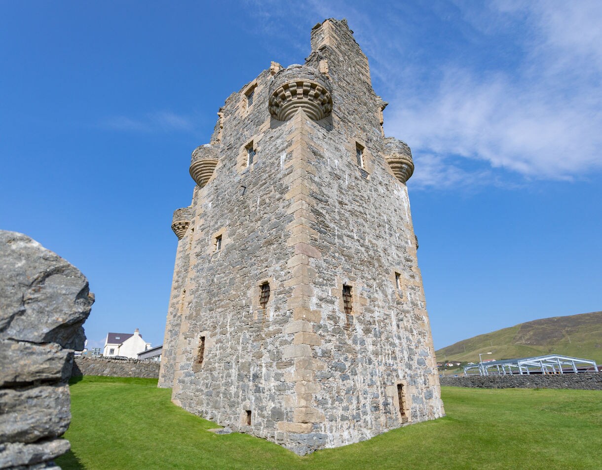 Stone tower of Scalloway Castle in Shetland, featuring thick walls, narrow windows and rounded corner turrets against a bright blue sky.