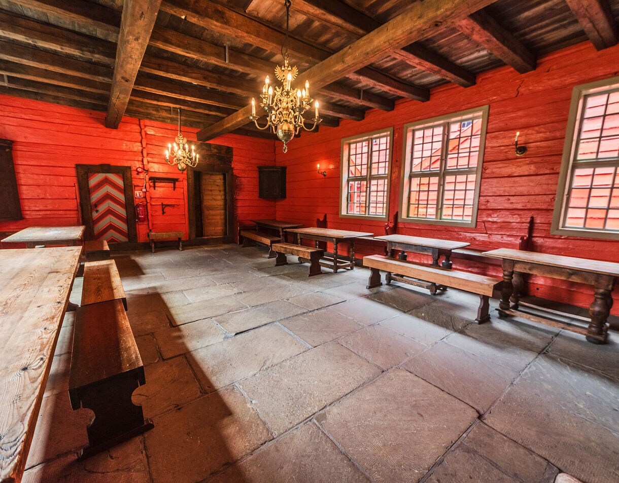 Interior of Schøtstuene in Bergen, featuring red wooden walls, stone flooring, long wooden tables with benches and ornate chandeliers.