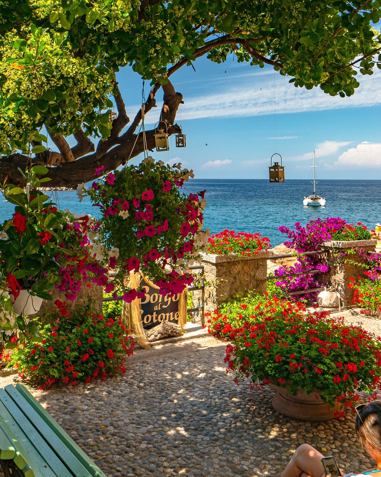 A colorful seaside terrace in Borgo al Cotone featuring hanging flower baskets, stone pathways and bright potted plants overlooking the calm blue water with a sailboat anchored offshore.