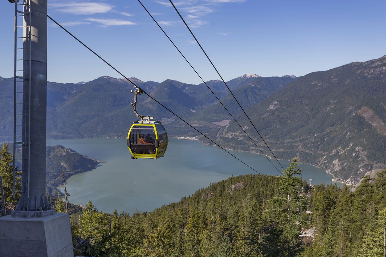 The Sea to Sky Gondola in Vancouver, BC, suspended high above a lush green forest, with stunning views of the blue waters of Howe Sound and the surrounding mountain ranges under a clear sky.
