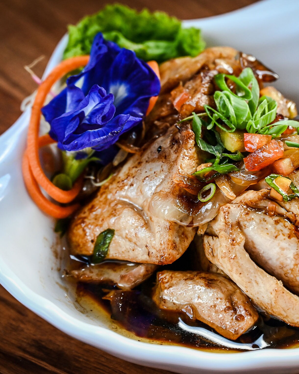 Bowl of glazed fish fillets in dark soy-based sauce, garnished with diced tomatoes, sliced green onions, carrot curls and a blue edible flower.