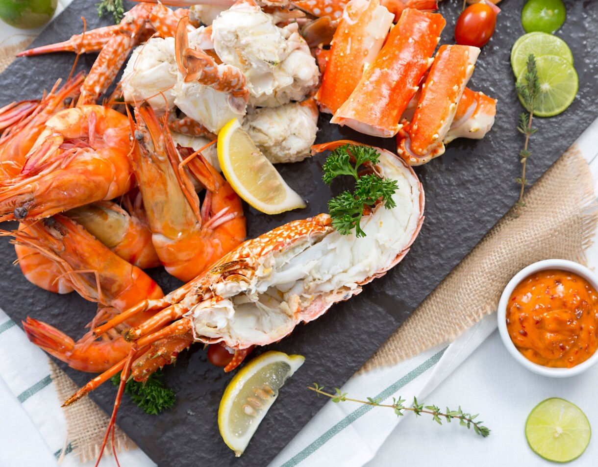 A seafood platter with cooked prawns, crab legs and a split lobster tail on a slate board, garnished with lemon wedges and parsley, with dipping sauces on the side.