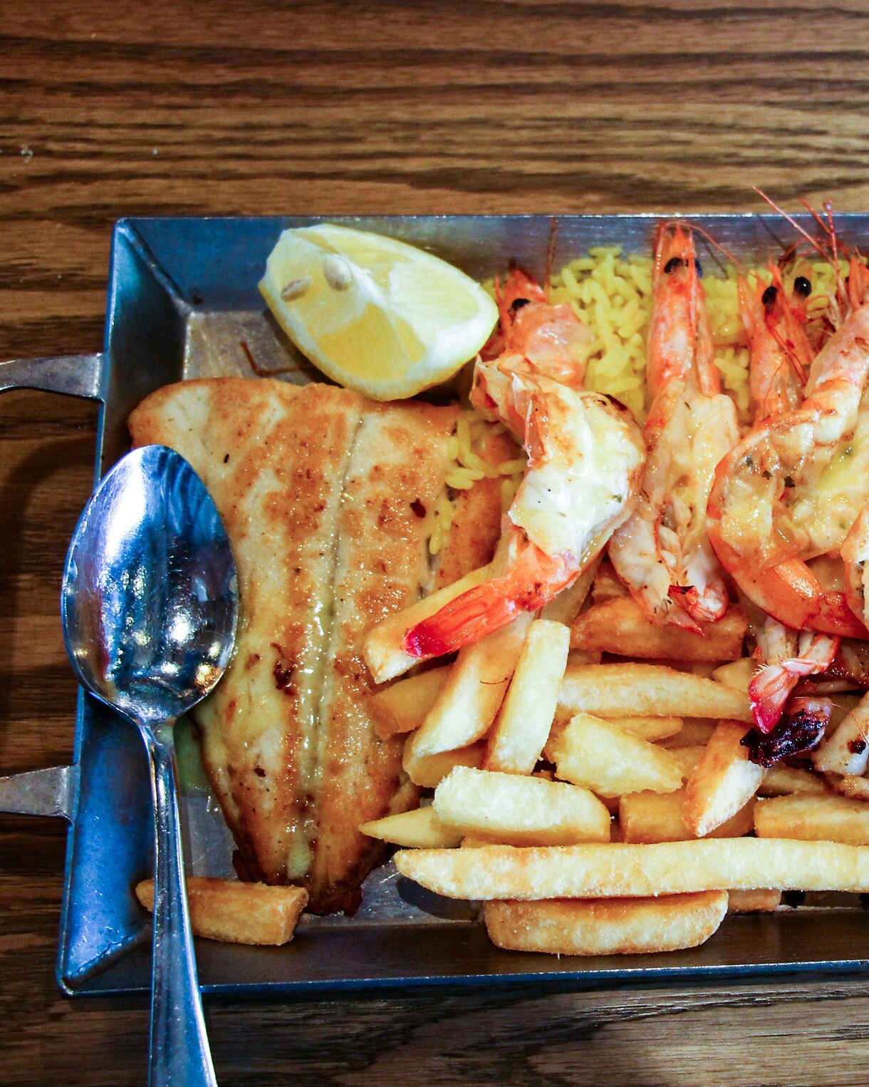A metal tray filled with assorted seafood including grilled fish, prawns, fried calamari and mussels in a creamy sauce, served with fries, rice and a lemon wedge.