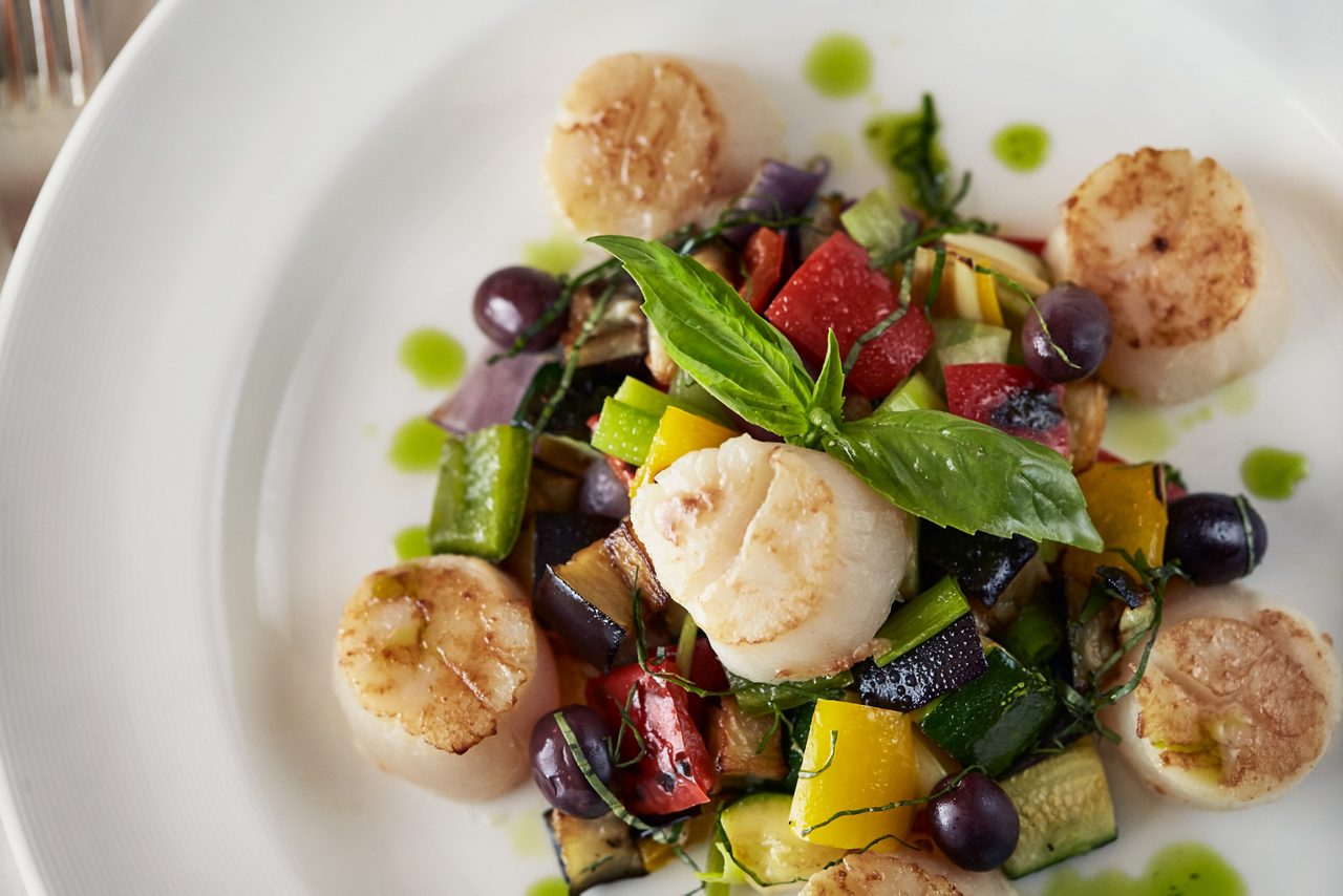 A close-up of seared scallops served with a vibrant mix of roasted vegetables, black olives, and fresh basil, drizzled with herb oil on a white plate. 