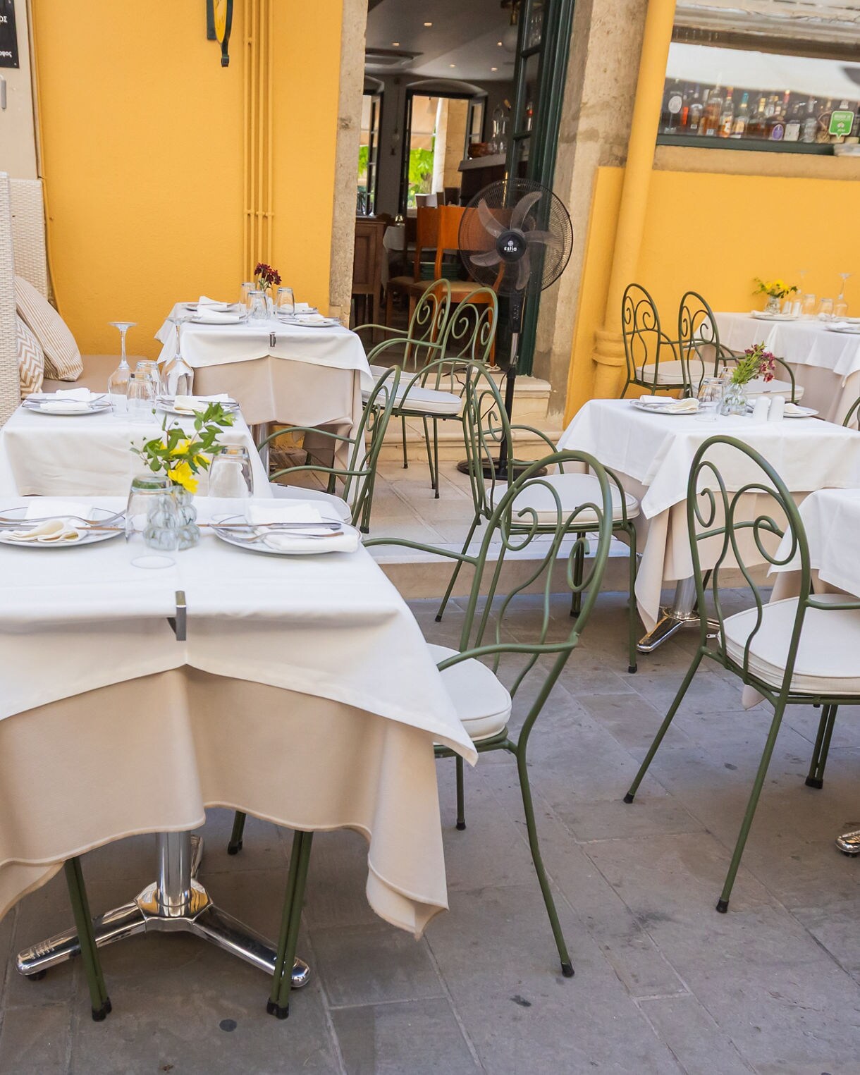 Outdoor café tables with white tablecloths and green wrought-iron chairs set against a warm yellow building wall.