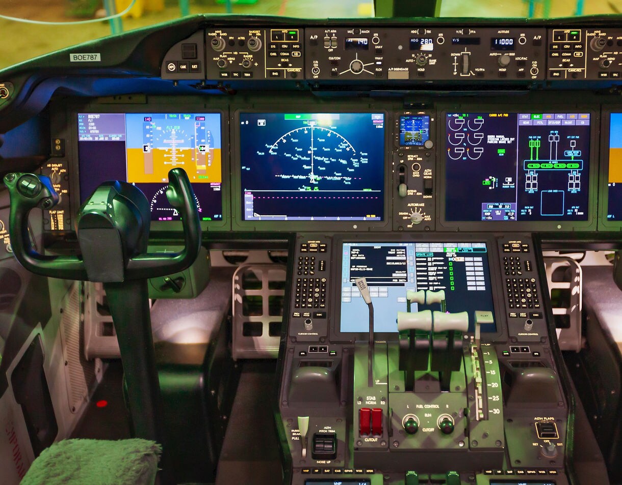 A detailed view of a Boeing aircraft cockpit showing multiple digital flight displays, control yokes, switches and throttle levers illuminated with bright instrument lights.