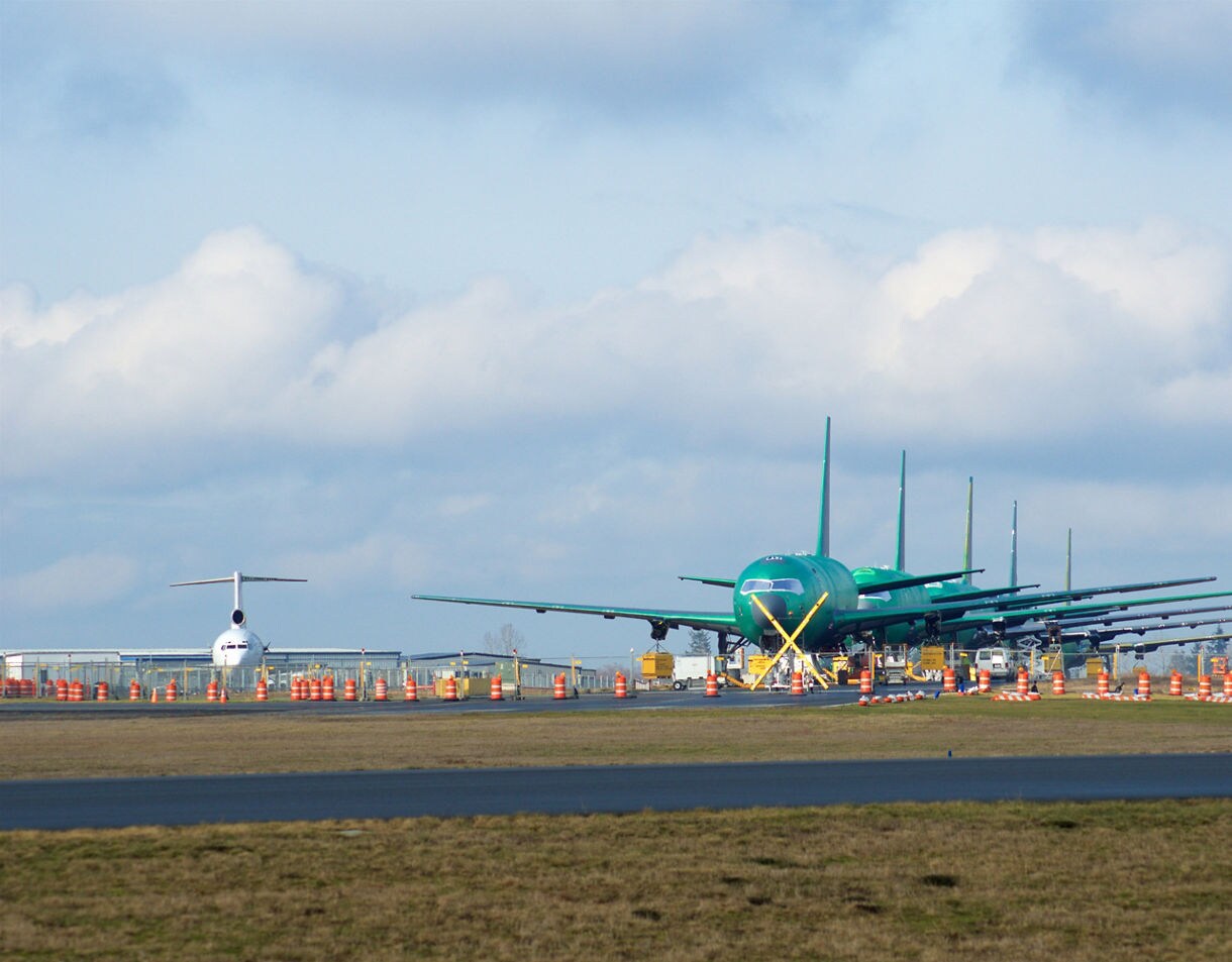 A row of unfinished Boeing aircraft with green protective coating parked on the tarmac, surrounded by equipment and orange safety cones under a cloudy sky.
