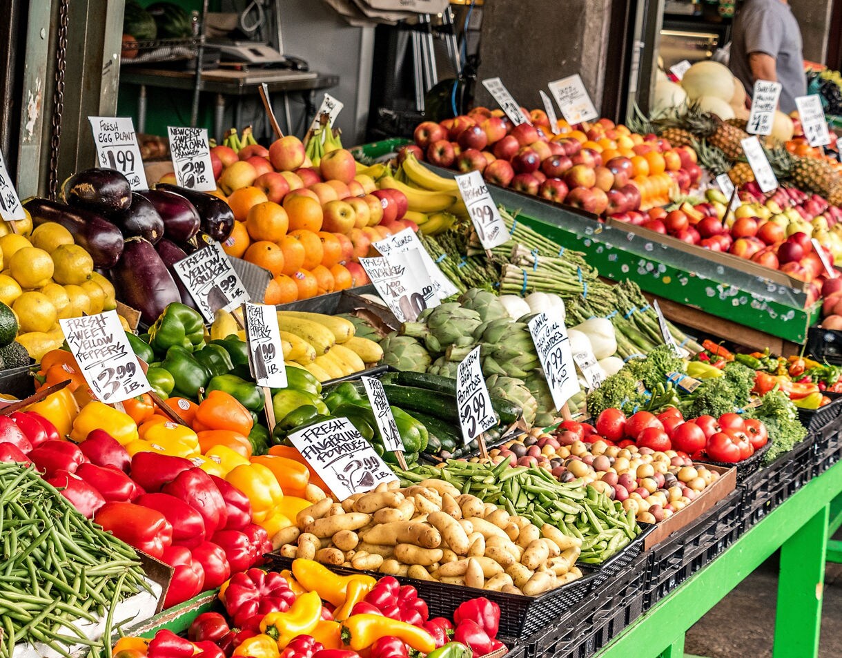 A colorful display of fruits and vegetables at Pike Place Market featuring tomatoes, avocados, lemons, peppers, beans, potatoes, apples and other fresh produce arranged on green tables with handwritten price signs.
