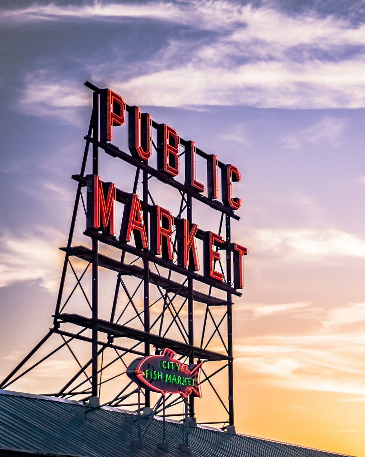A view of the neon “Public Market” sign at Pike Place Market in Seattle silhouetted against a colorful sunset sky with soft clouds and warm light.