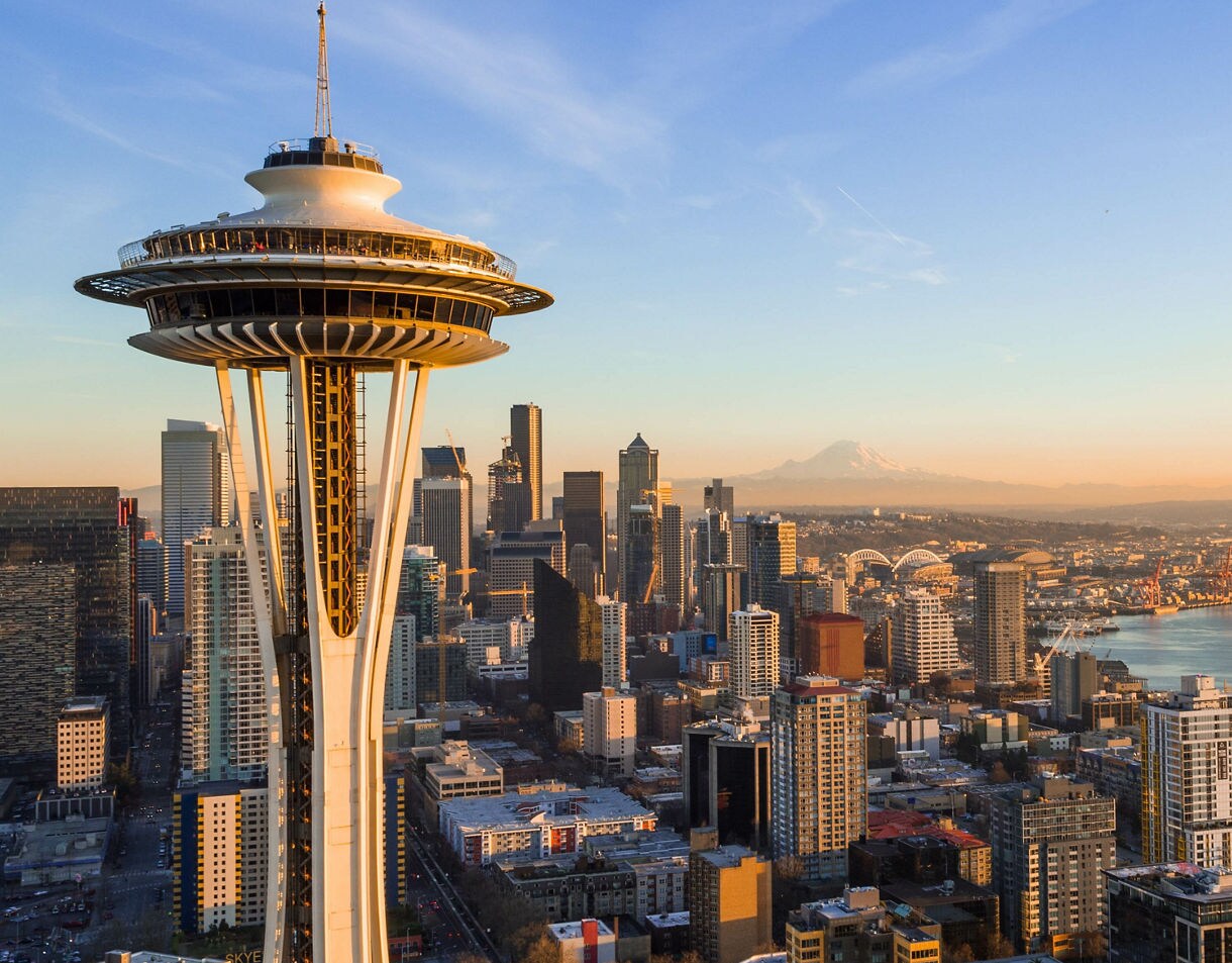 An aerial view of Seattle featuring the Space Needle in the foreground, high-rise buildings across downtown, the waterfront and distant Mount Rainier under a warm sunrise sky.