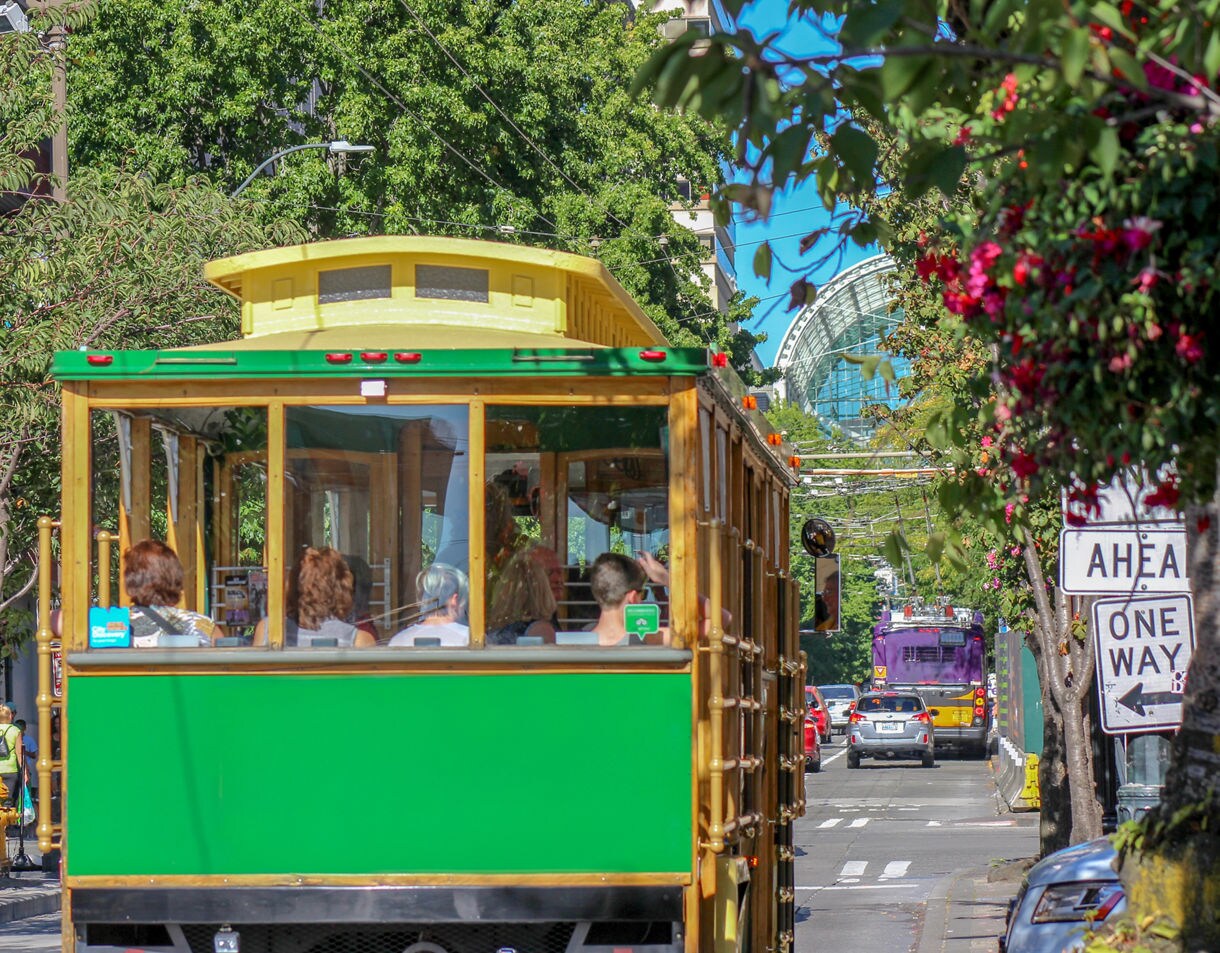 A colorful trolley drives along a busy Seattle street with trees, signage, and a glass-domed building visible in the distance.