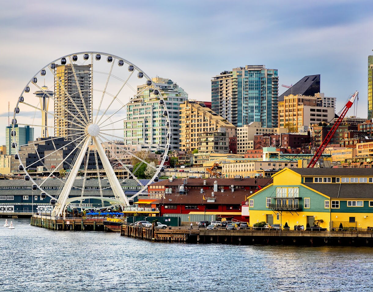 Seattle waterfront with the Great Wheel in front of downtown high-rises and colorful pier buildings along the bay.