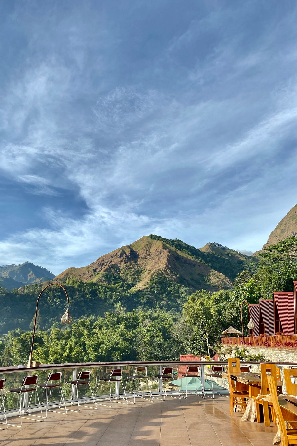 Outdoor terrace with wooden tables and tall chairs overlooking lush green hills and rugged mountain peaks under a bright blue sky with wispy clouds.