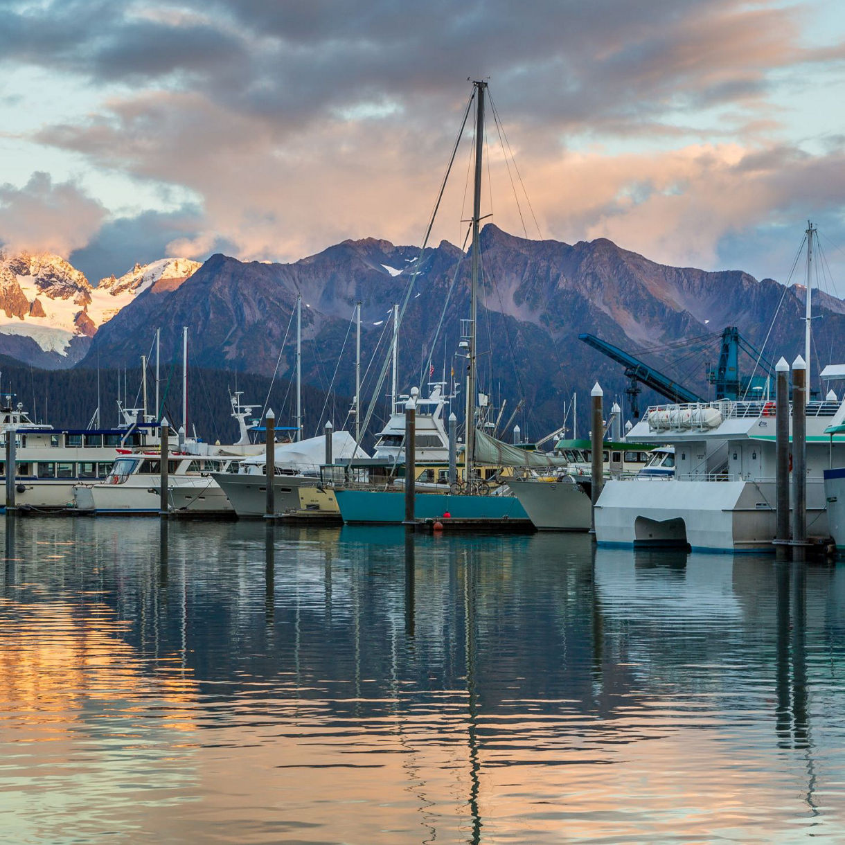 A serene marina with boats docked, reflecting mountain peaks and sunset hues against a backdrop of snow-capped mountains.