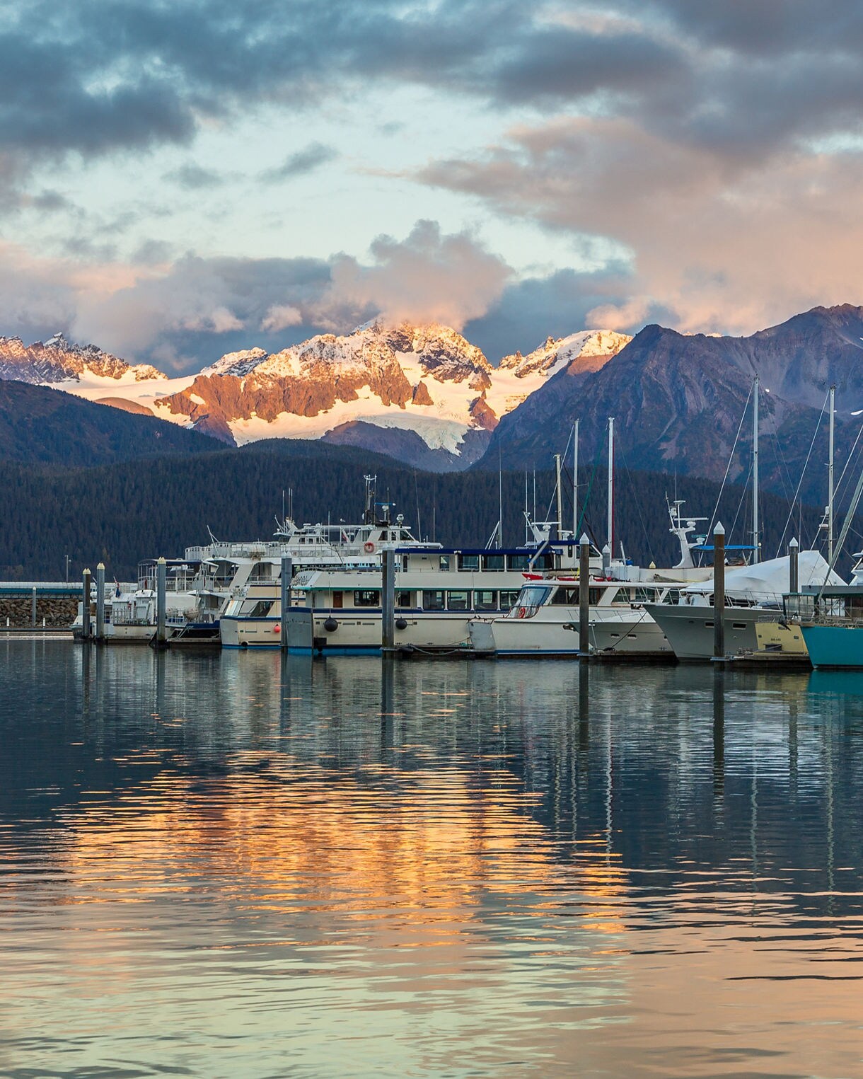 Boats docked at Seward Harbor in Alaska with golden sunset light reflecting off the water and snow-covered mountains glowing in the background under a cloudy sky.