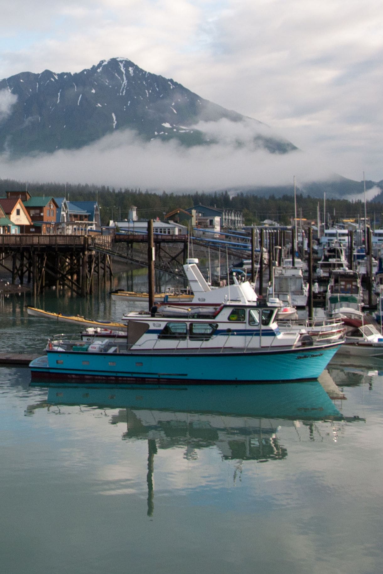 A peaceful view of Seward Harbor in Alaska, featuring boats docked on calm waters, waterfront buildings, and misty mountains in the background.