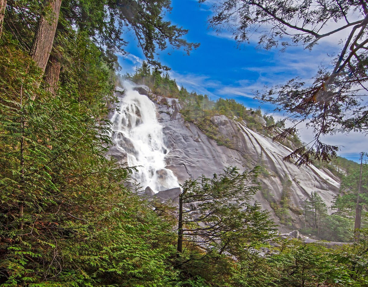 Waterfall cascading down granite cliffs surrounded by lush evergreen forest under a bright blue sky.