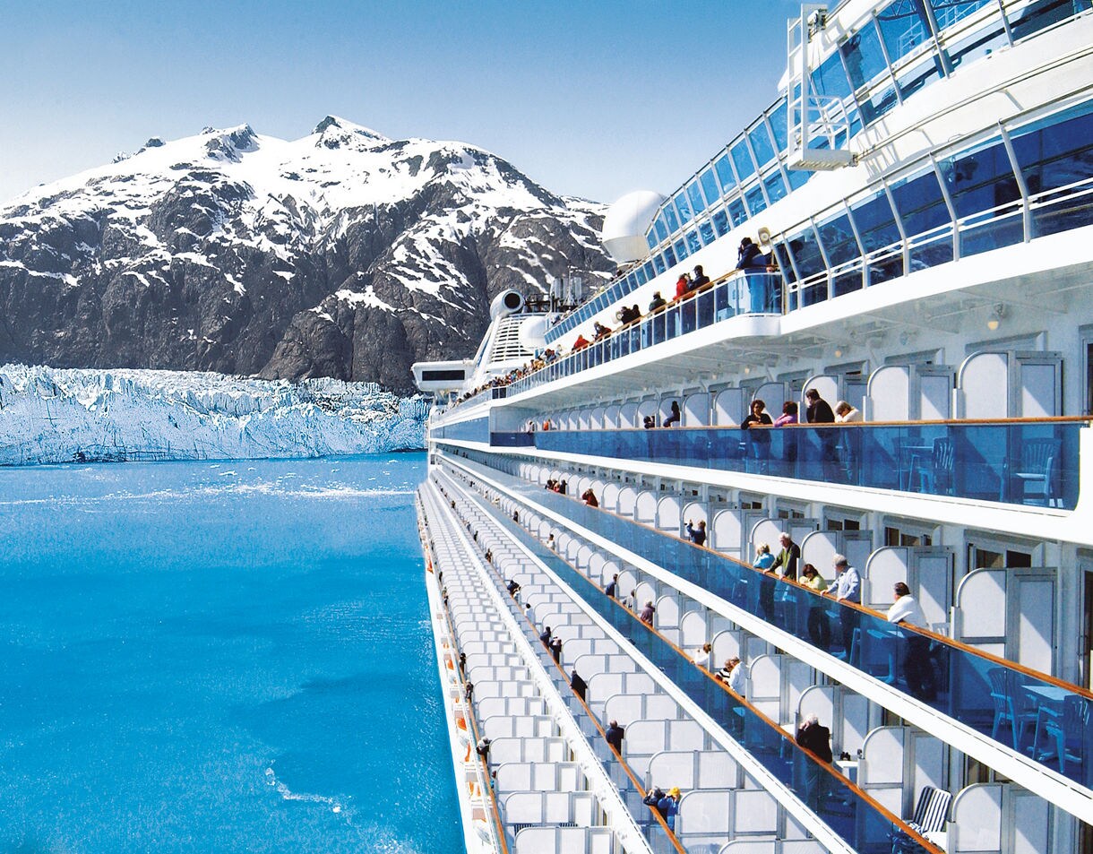 A large cruise ship sails near a stunning icy mountain range surrounded by bright blue waters. Passengers are visible enjoying the view from the ship's balconies. The scene captures the serene beauty of a cold, remote destination with clear skies and vibrant colors.