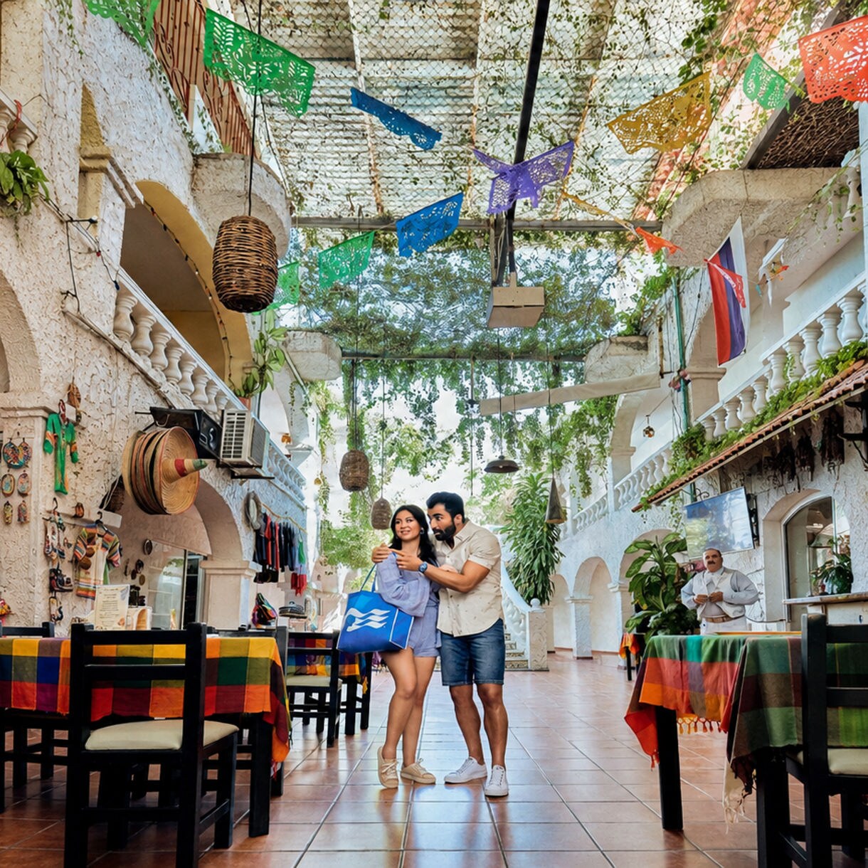 A young couple stands together in a vibrant open-air courtyard, appearing to explore or choose a table at a restaurant. The setting features rustic stone walls, hanging plants, woven baskets, and multicolored textiles covering the tables. Festive paper decorations are strung overhead, adding to the lively atmosphere. The scene suggests a warm, welcoming destination with cultural decor and outdoor dining.
