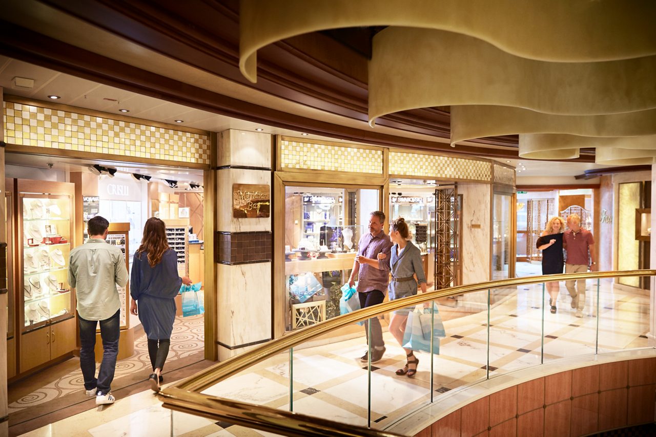 Two couples browse illuminated retail displays while walking through an onboard shopping promenade.