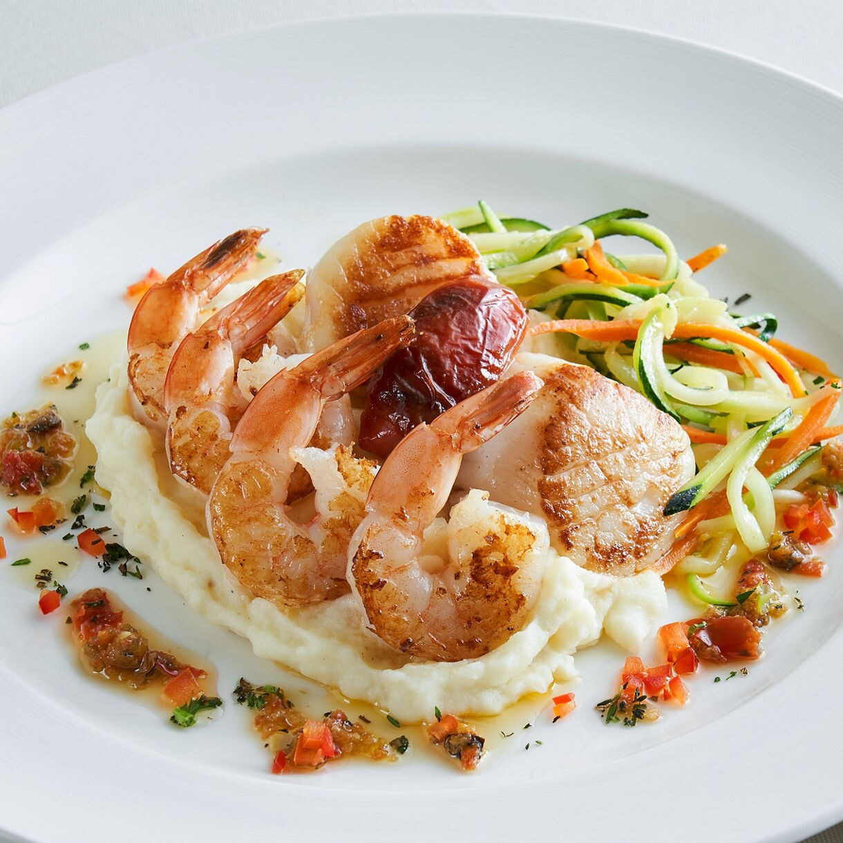 An elegant dish featuring succulent shrimp arrayed around a grilled scallop and a plump, roasted tomato, served atop creamy mashed potatoes, accompanied by a garnish of spiralized zucchini and carrot.