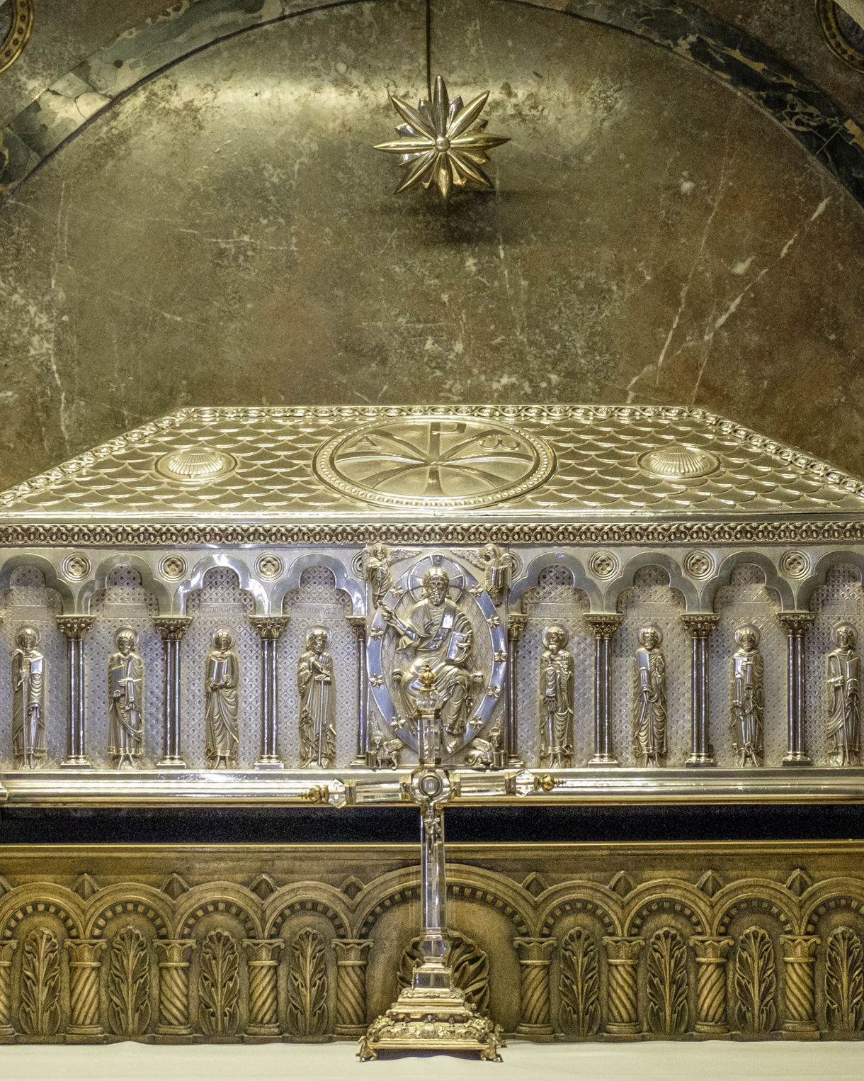 A silver reliquary adorned with carved saints and ornate detailing, displayed in a stone crypt beneath an arched ceiling with a small star-like ornament above.