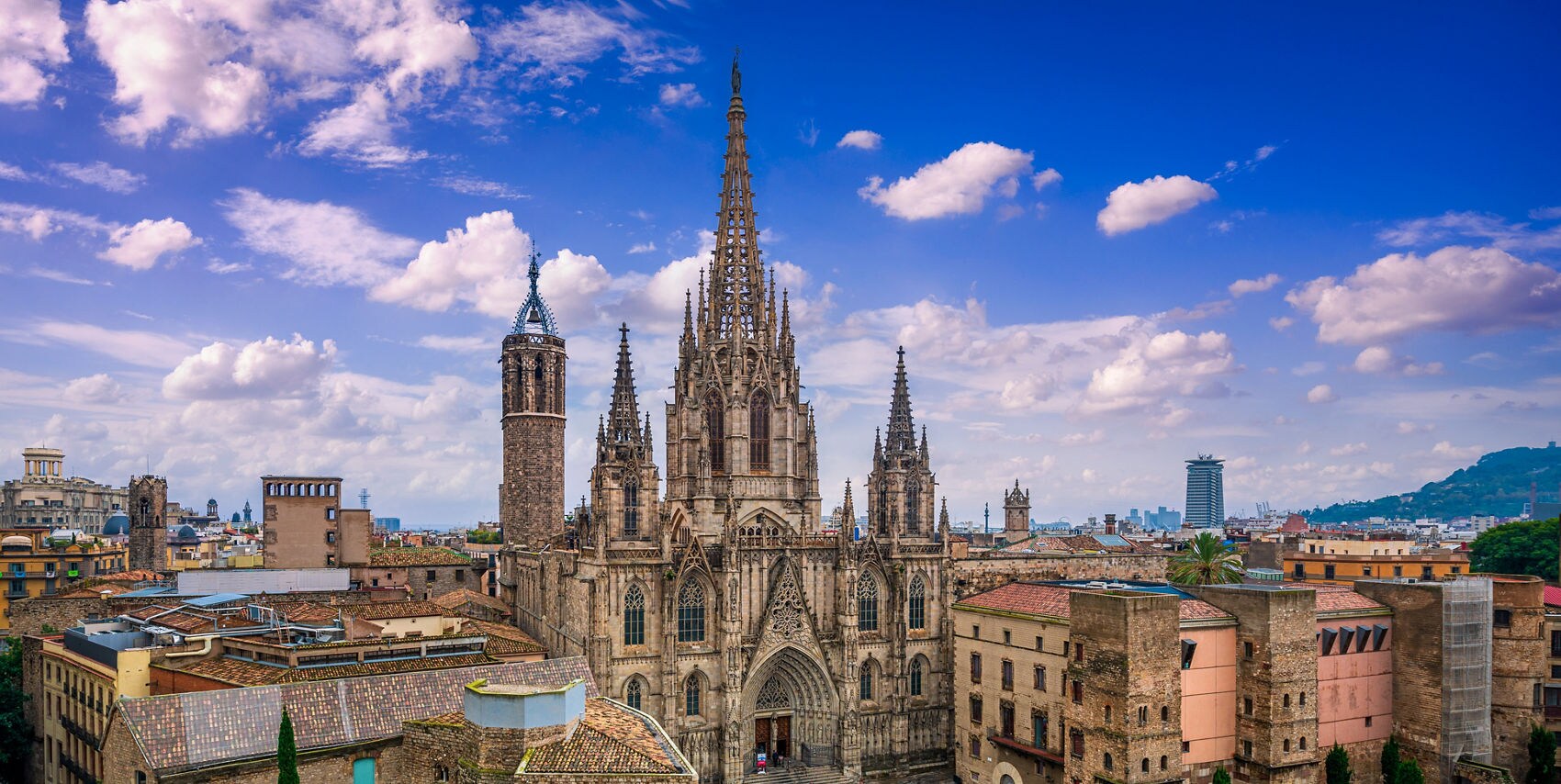 Aerial view of the Gothic Cathedral of Santa Eulàlia in Barcelona's Gothic Quarter, its intricate spires and ornate facade dominating a busy stone plaza surrounded by ancient buildings and the city skyline beyond.