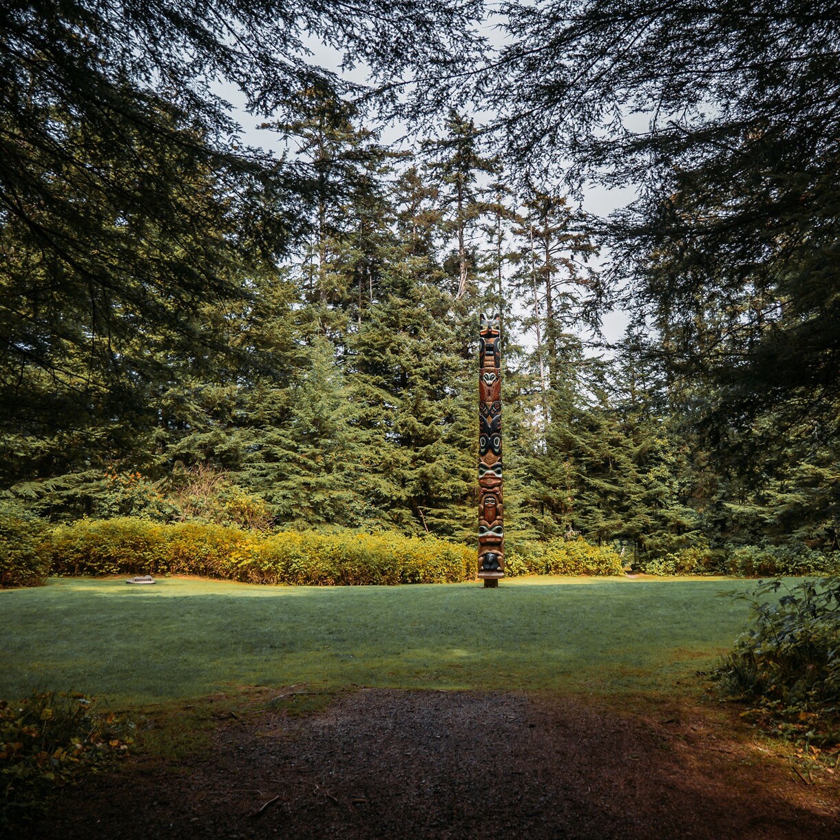 A single tall totem pole stands in a clearing surrounded by lush forest at Sitka National Historical Park, with sunlight filtering through dense evergreen trees.