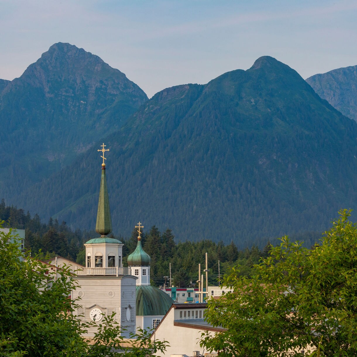 St. Michael’s Cathedral with its green domes and golden crosses stands in front of forested mountains in Sitka, Alaska under a clear blue sky.