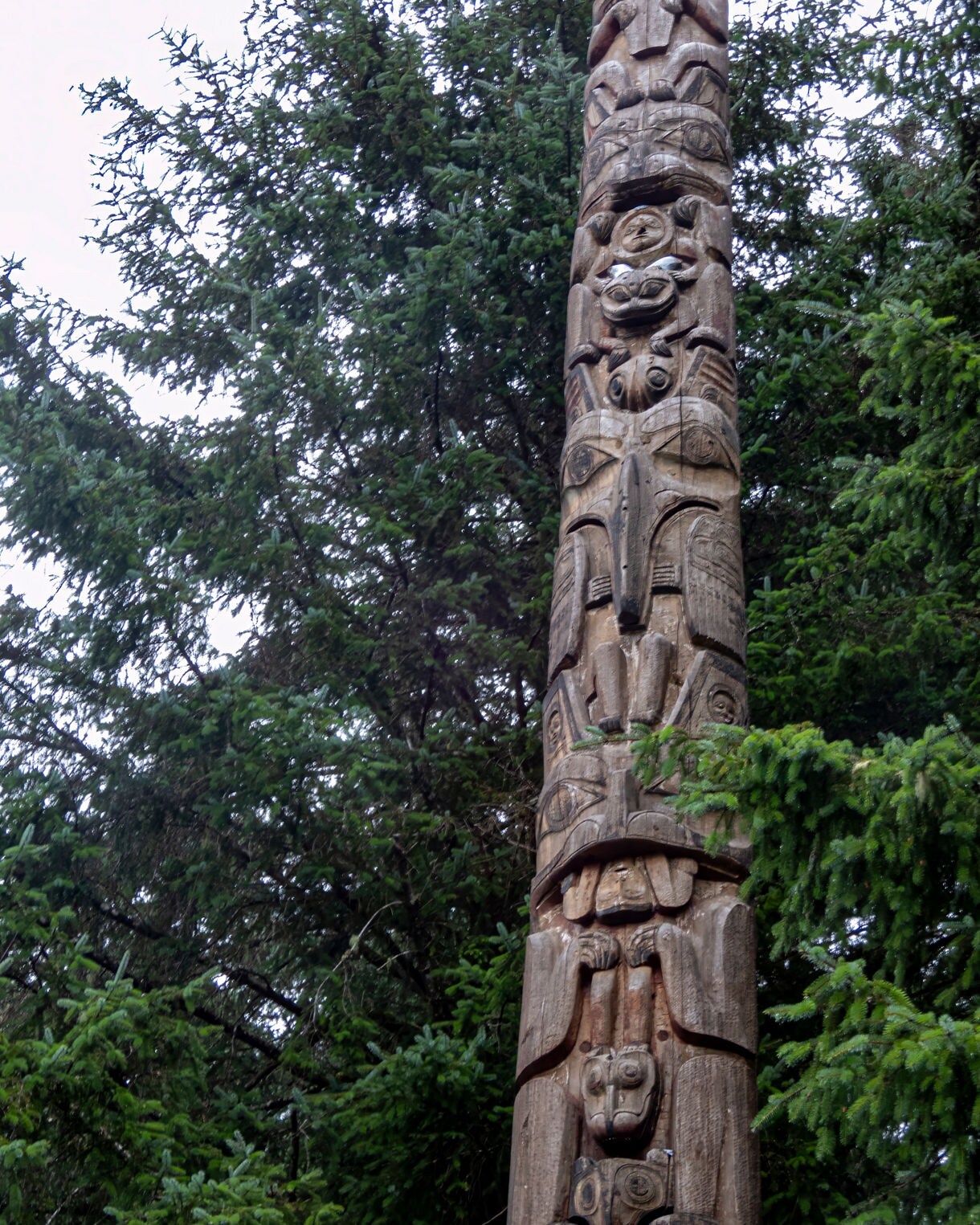 Close-up view of a traditional wooden totem pole carved with animal and human figures, set against dense evergreen trees in Sitka, Alaska.