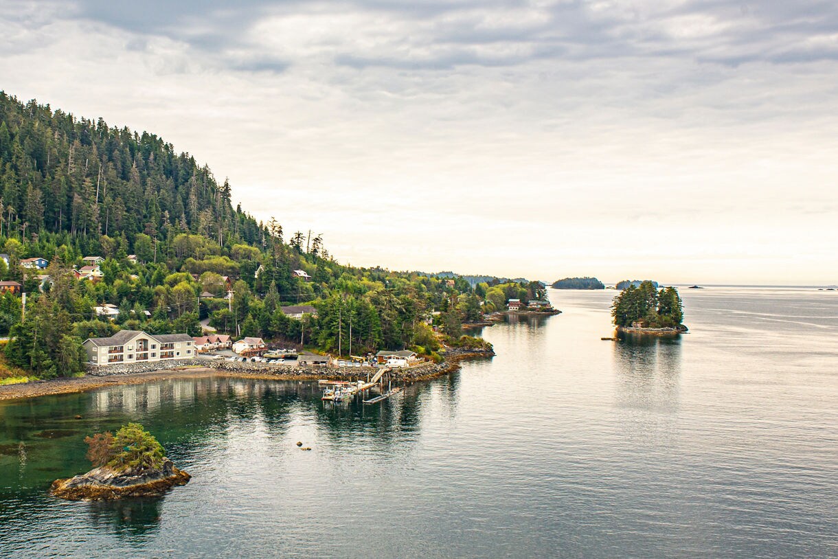 Aerial view of Sitka, Alaska featuring coastal homes, dense green forest, a small harbor with boats and tiny tree-covered islands scattered in the calm ocean.