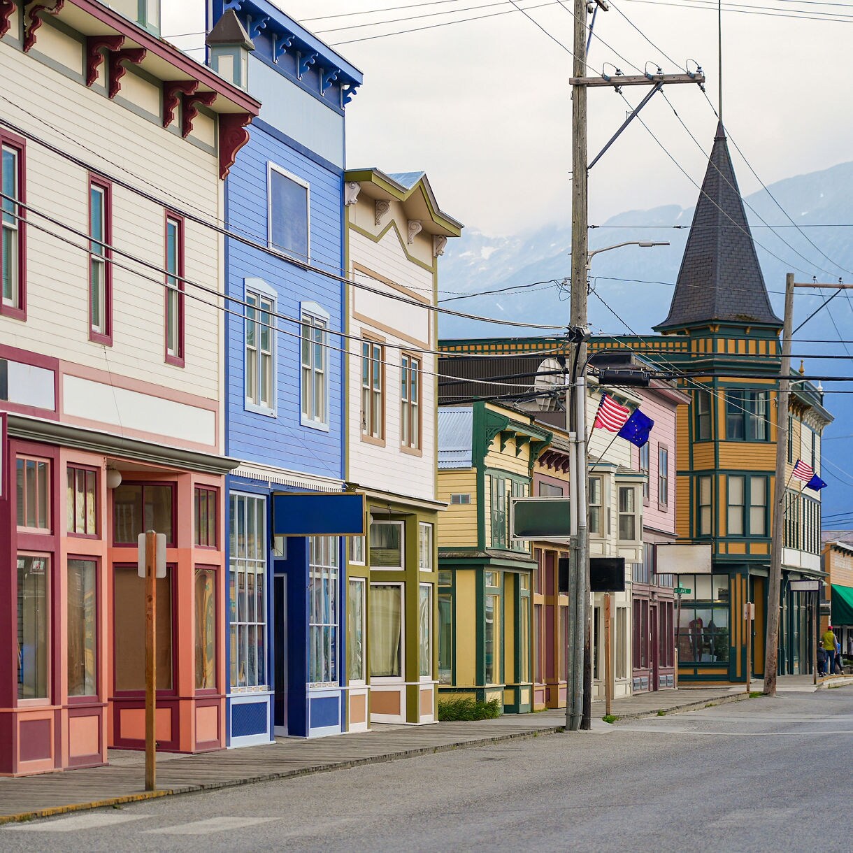 A row of colorful historic buildings lining a street in Skagway, Alaska, with flags fluttering in the breeze and a cruise ship visible in the distance.