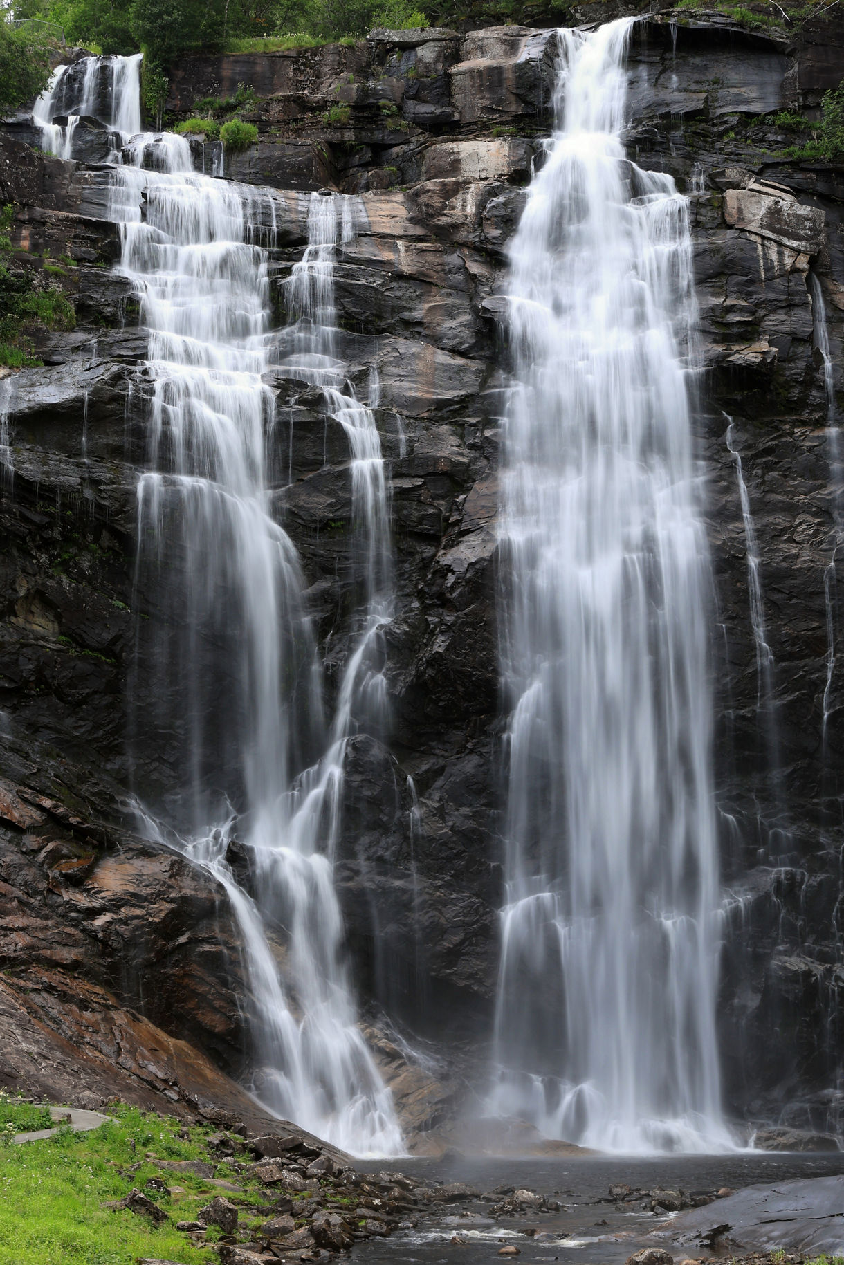 Skjervefossen waterfall in Norway, featuring twin streams plunging down dark cliffs into a misty basin surrounded by greenery.