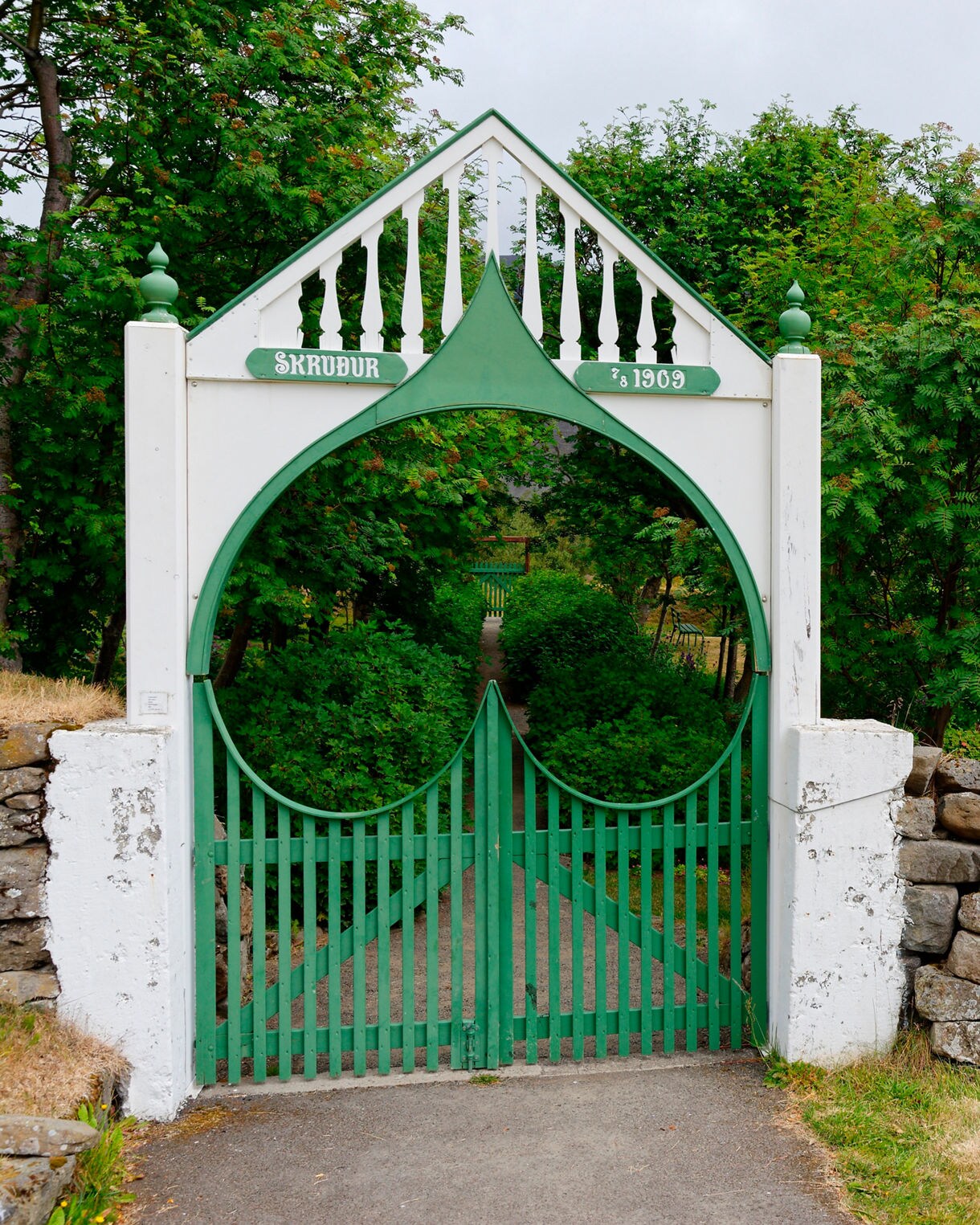Green and white gate at the entrance to Skrúður Botanical Garden, framed by stone walls, trees and mountain backdrop.