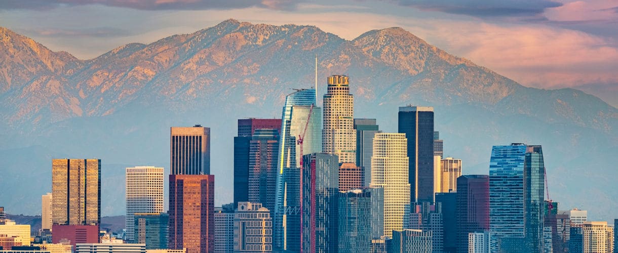 Downtown Los Angeles skyline with tall skyscrapers, framed by the San Gabriel Mountains and a colorful sunset sky.