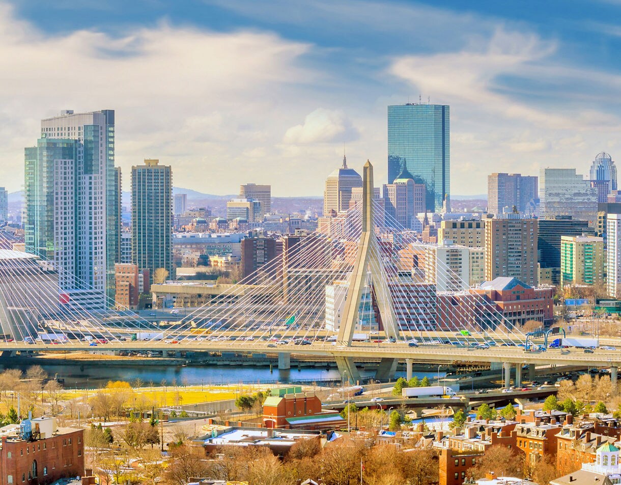 Wide view of the Boston skyline featuring the cable-stayed Zakim Bridge, high-rise buildings and the Charles River under a partly cloudy sky.
