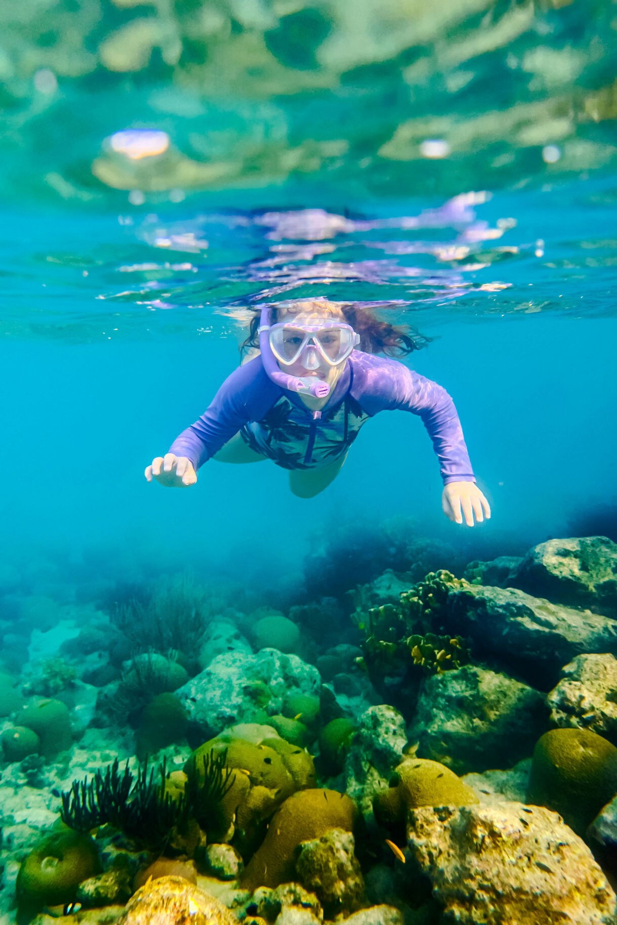 A snorkeler in a blue rash guard and fins glides through clear turquoise water, swimming next to a sea turtle with a patterned shell near the ocean floor.