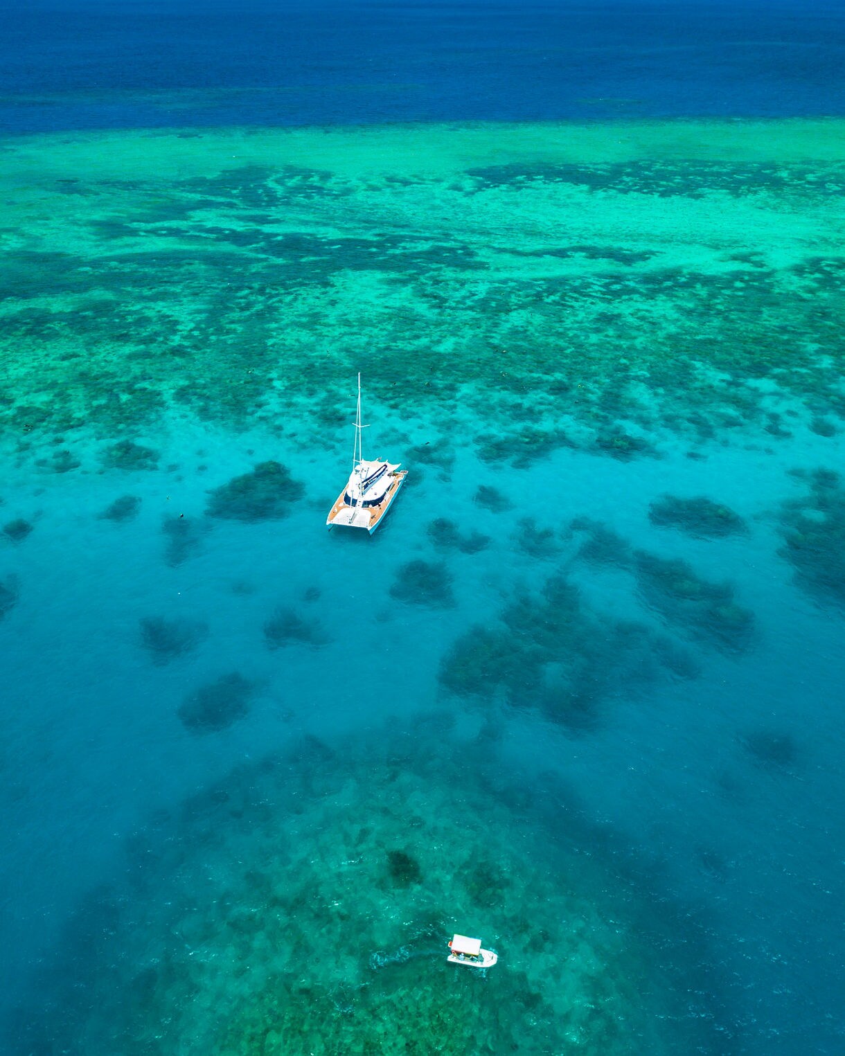 A catamaran anchored over clear turquoise water with coral patches visible below and a small snorkel boat nearby.