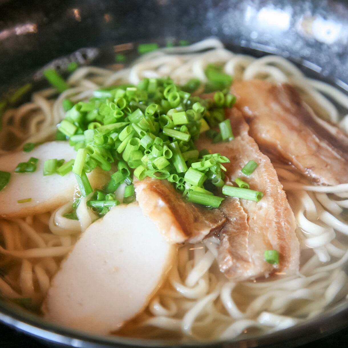 Close-up of a bowl of Okinawan soki soba featuring thick noodles in clear broth topped with sliced pork, fish cake and a generous handful of chopped green onions.