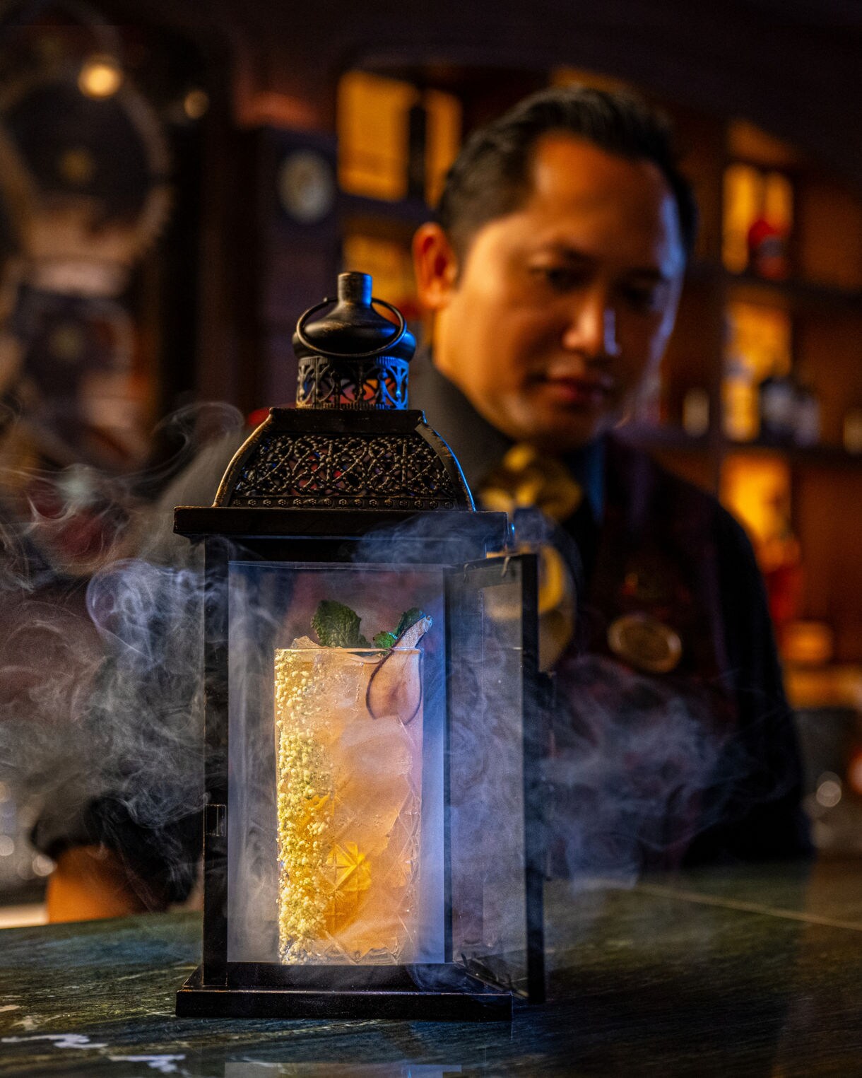 A smoky lantern containing a glowing cocktail sits on a bar counter while a bartender observes in the background.
