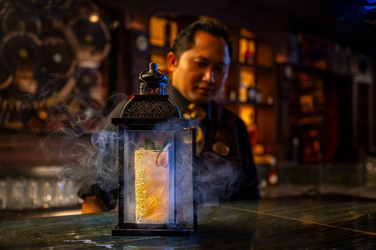 A bartender holding an illuminated vintage-style lantern in a dimly lit bar setting with bottles visible in the background.